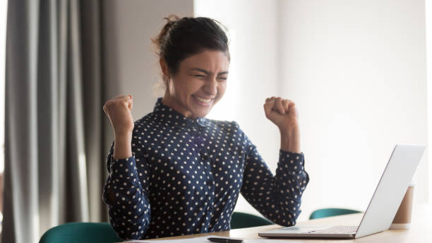 Happy indian woman office worker feeling excitement raising fists celebrates career ladder promotion or reward, businesswoman sitting at desk receive online news, great results successful work concept